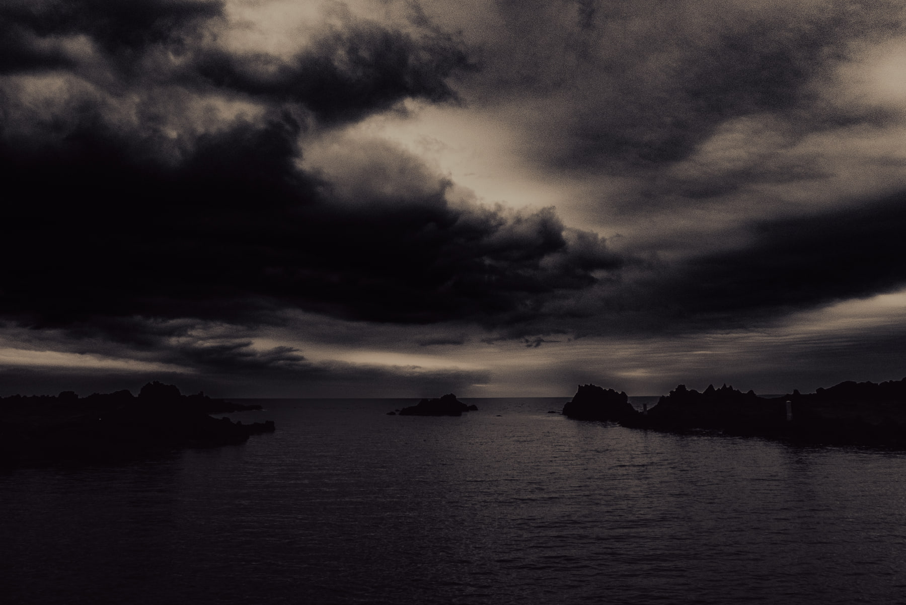 Heavy dark cloud presses down over a flat sea and low rock outcrops.