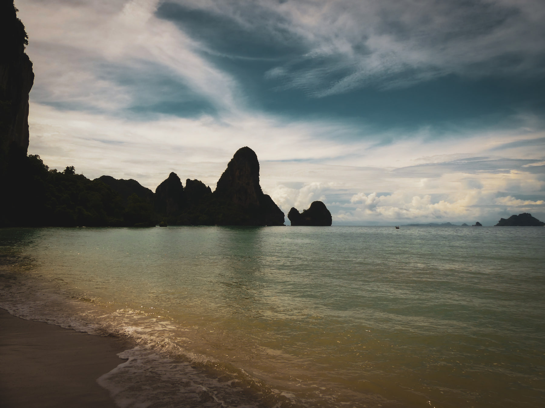 Limestone cliffs stand over a calm bay under heavy cloud, with a pale beach in the foreground.