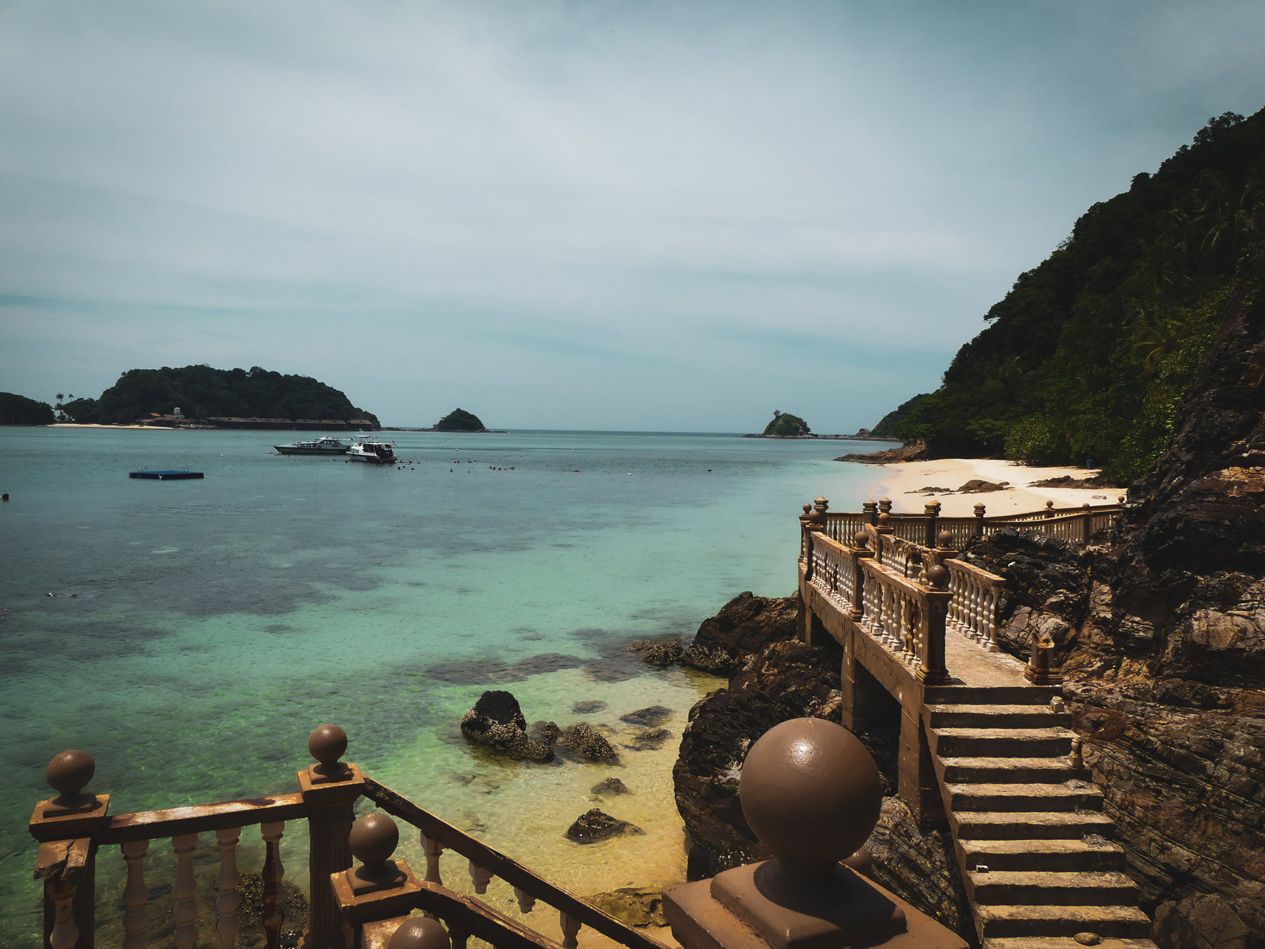 A balustraded stone stair drops to rocks and a shallow bay, with a moored boat, a small beach and low islands beyond.