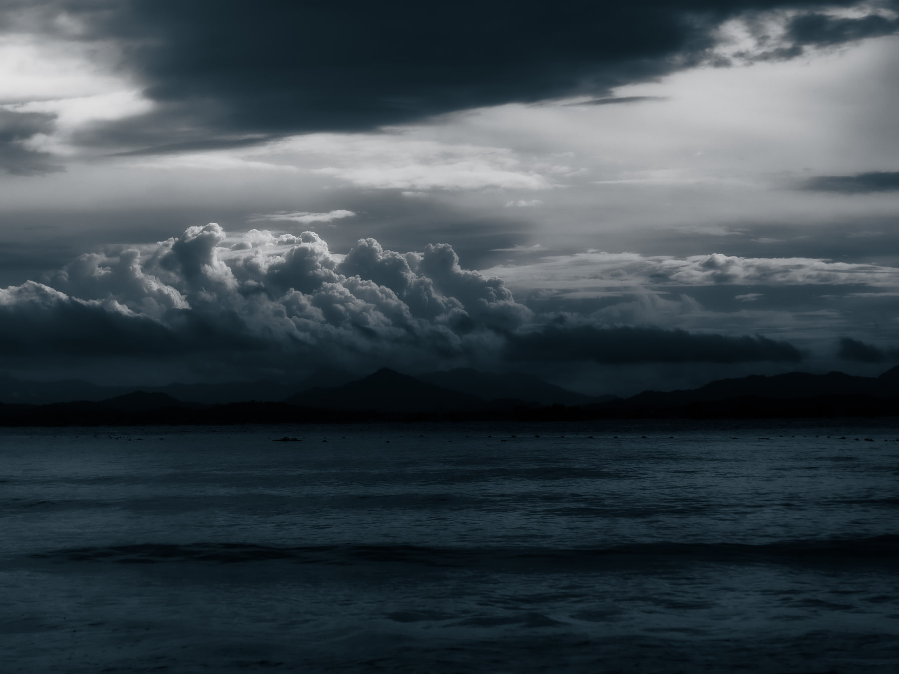 A bank of cumulus catches the last light over a dark sea, with mountains low on the far shore under heavy cloud.
