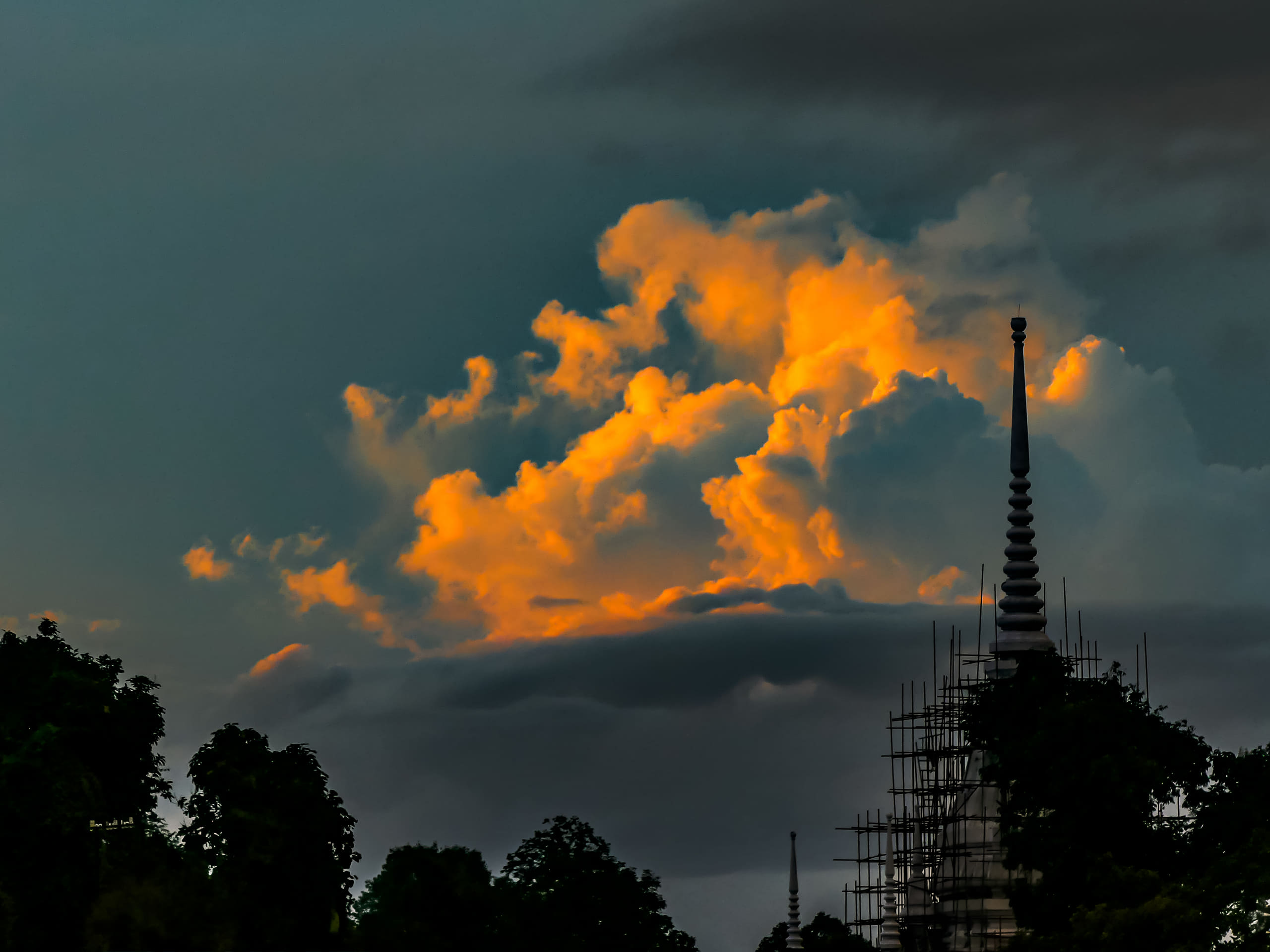 A bank of cloud lit gold by low sun stands behind the tiered spire of a temple wrapped in bamboo scaffolding.