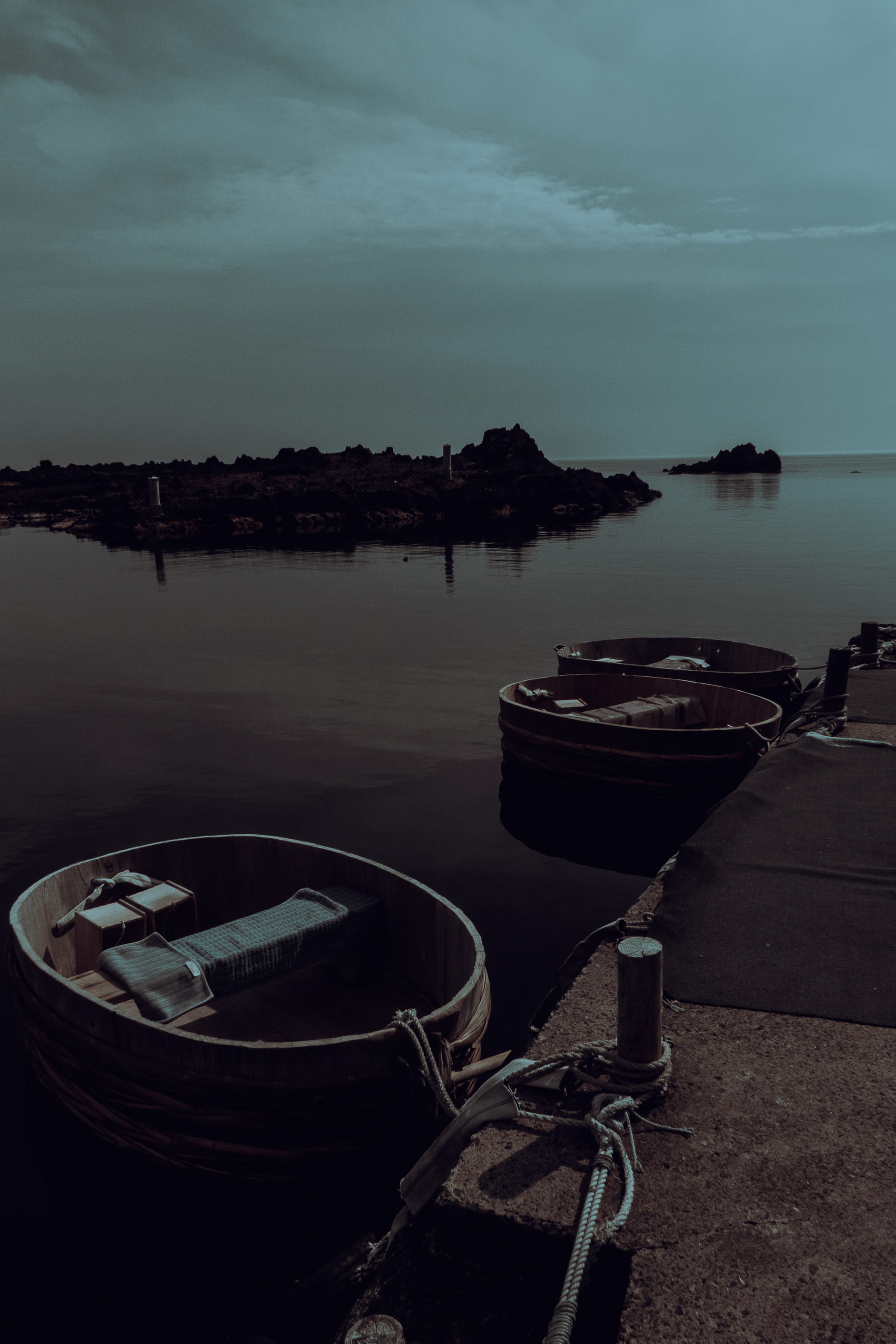 Three round wooden boats lie moored along a stone quay beneath a low grey sky, with black rocks across the water behind them.