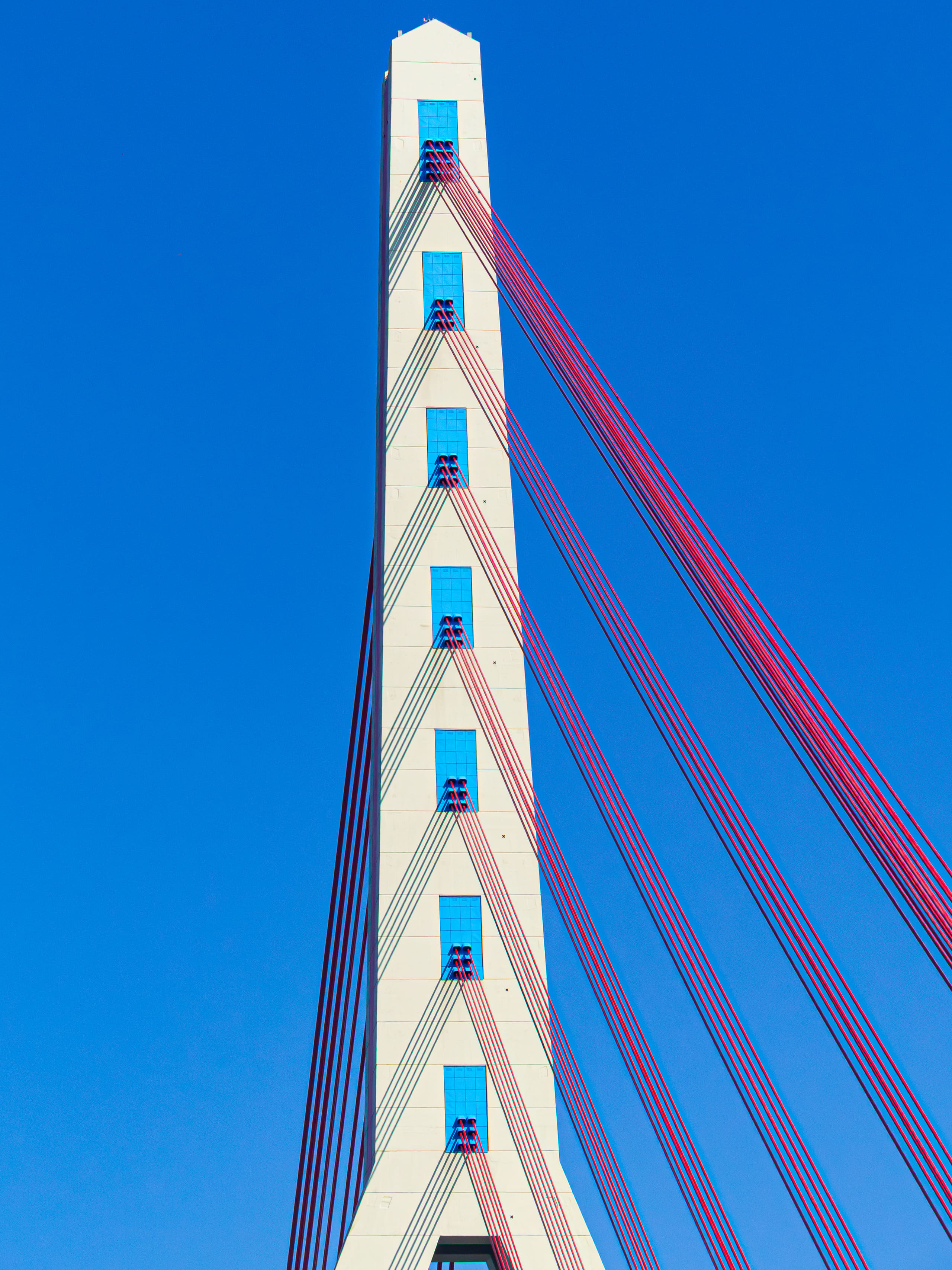 Red cables run down from the white pylon of a bridge into deep blue sky.