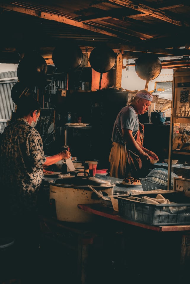 Two cooks work in a dim open kitchen hung with pans.