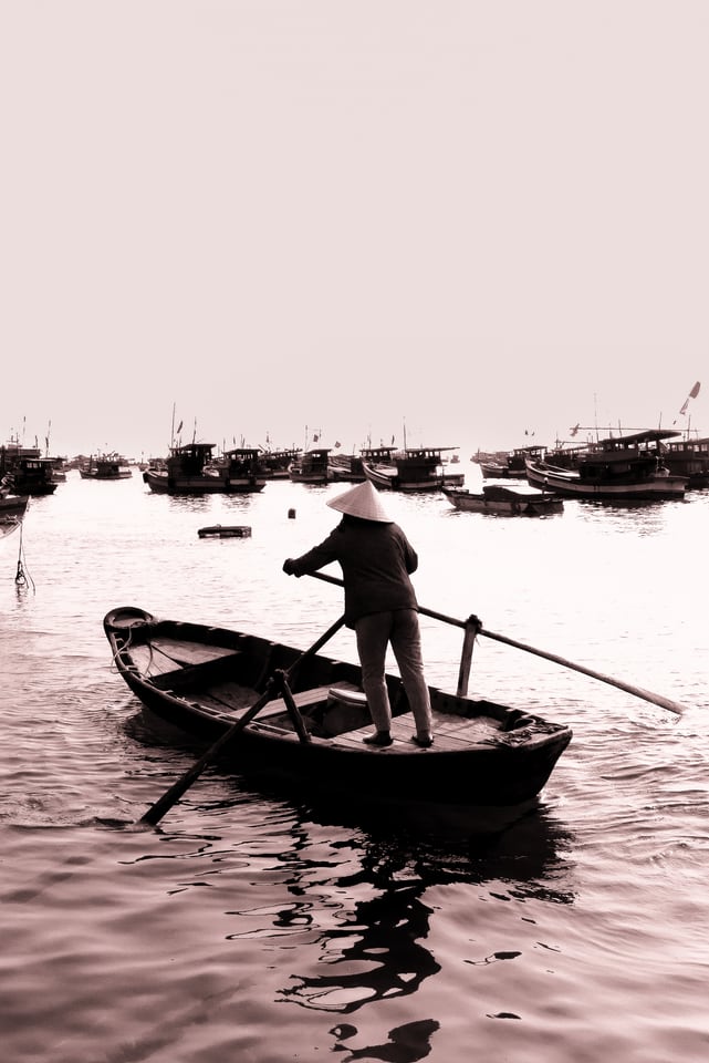 A person in a conical hat rows a small boat past a fleet at anchor.