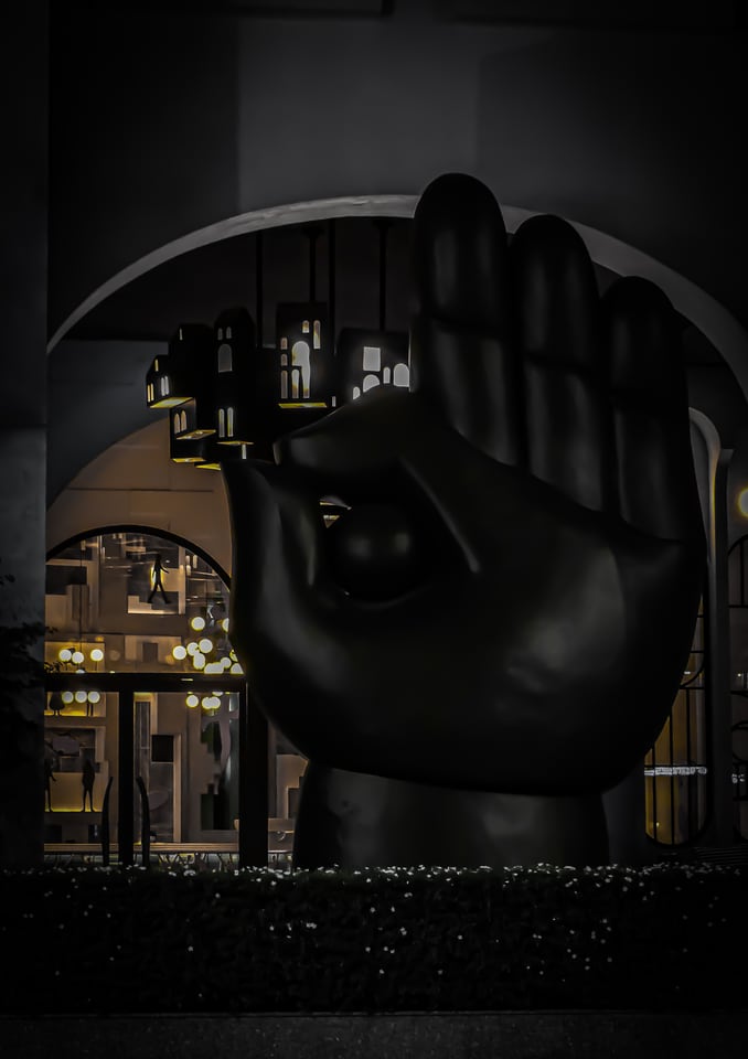 A large dark sculpture of a hand stands before an arcade at night.
