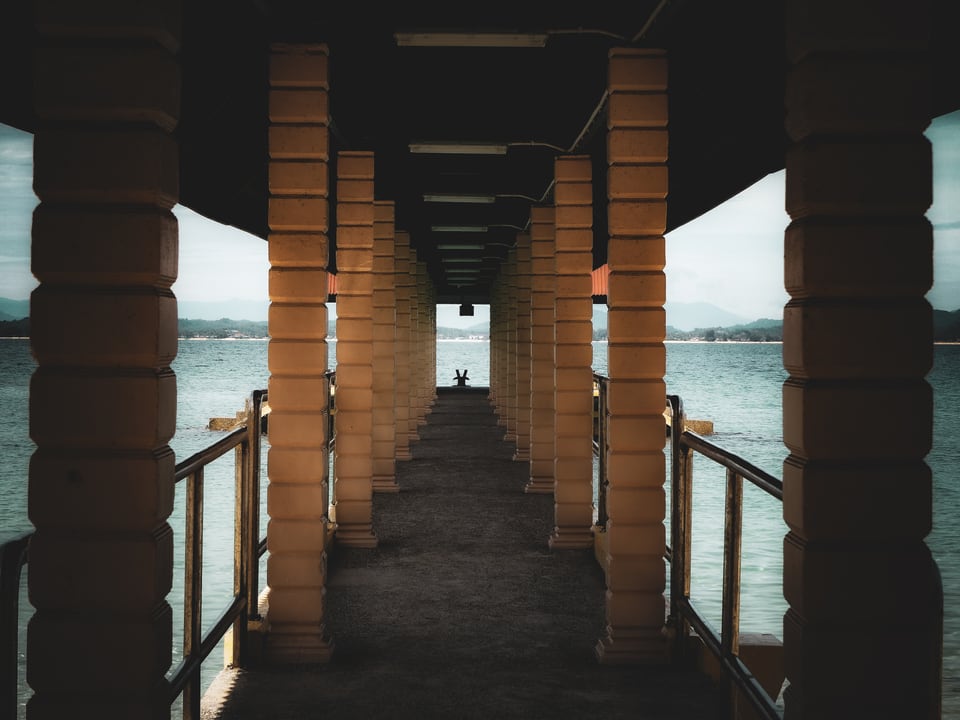 A covered walkway runs between heavy square columns towards open sea, with a single distant figure at the far end.