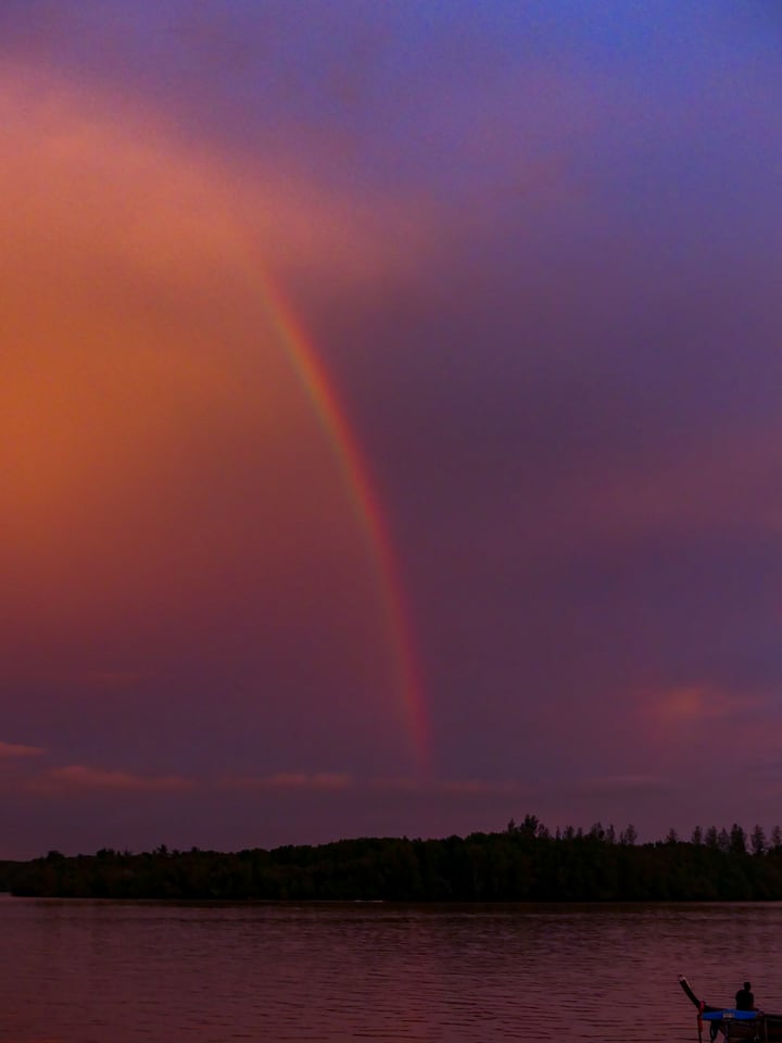 A rainbow arches over a dark treeline and still water.