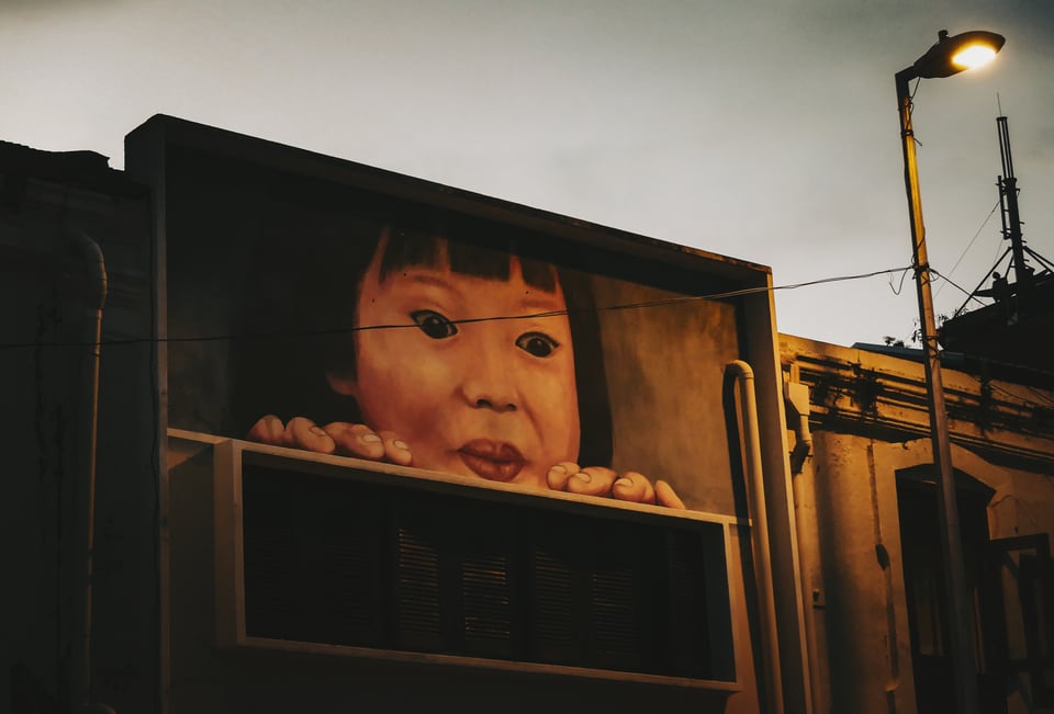 A mural of a child's face peers over a painted ledge on a building.