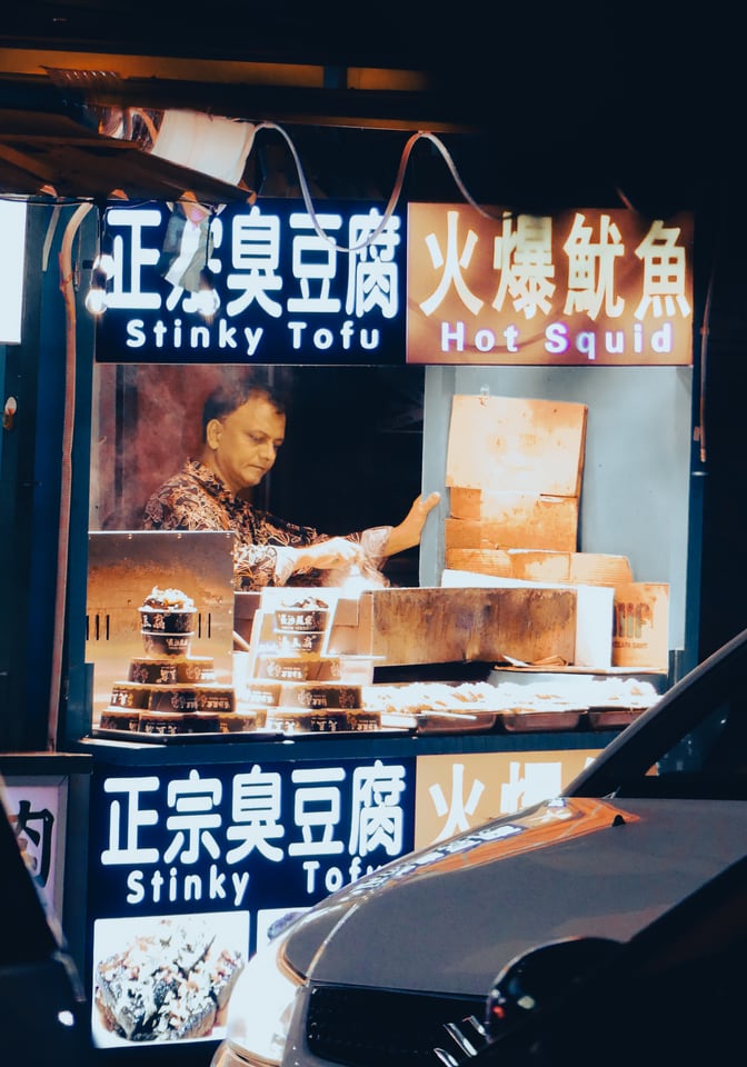 A vendor works behind the lit signs of a street food stall.