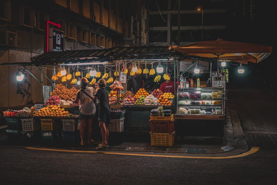 A lit fruit stall stands on a dark street corner at night.