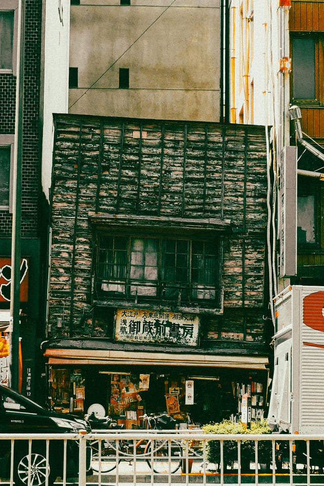 A weathered green timber shopfront with faded signboards is wedged between two much larger modern buildings.