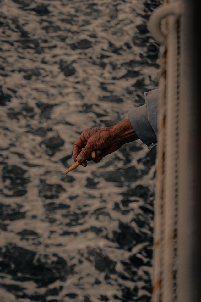 A weathered hand reaches out past a ship’s rail, holding something small above moving water.