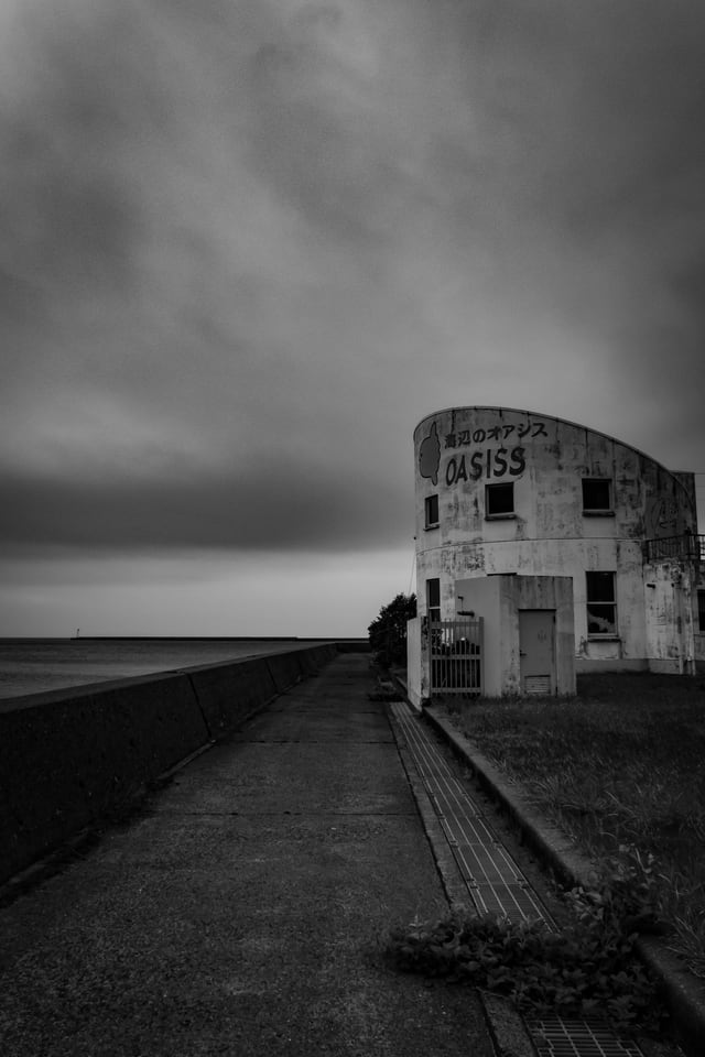 A single building stands at the end of a seawall road under heavy cloud.