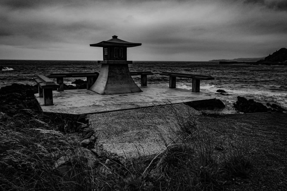 A stone shrine stands on a shingle shore beneath heavy cloud.