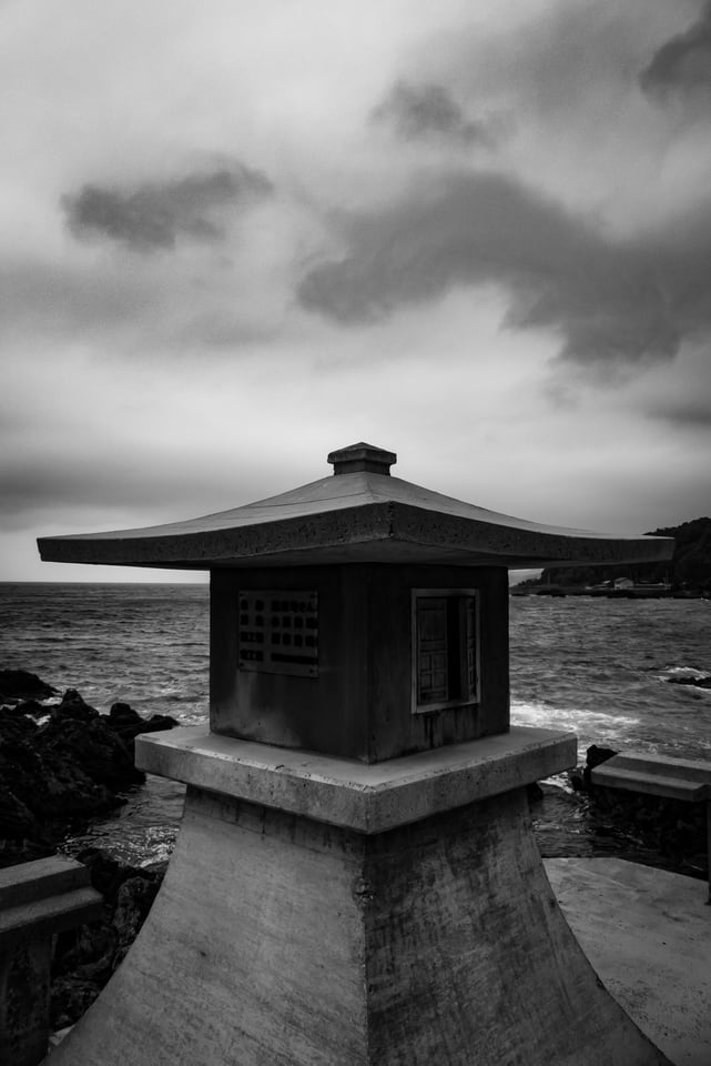 A small stone shrine lantern stands at the end of a concrete path above the sea.