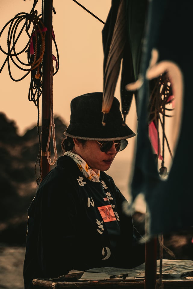 A performer in a dark hat waits beneath festival ropes.