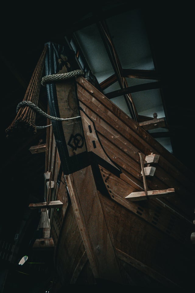 The timber prow of a wooden boat, roped and cleated, seen close and from below inside a shed.