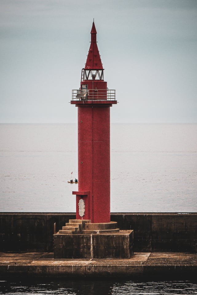 A red lighthouse stands at the end of a breakwater under a pale sky.