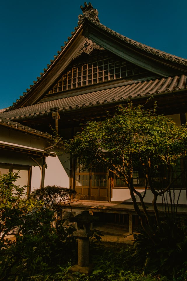 A temple hall with a heavy tiled roof stands among trees under a blue sky.