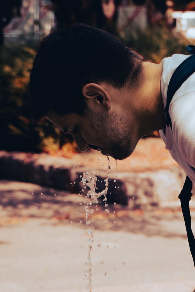 A man bends to drink, water falling from his mouth in low sun.