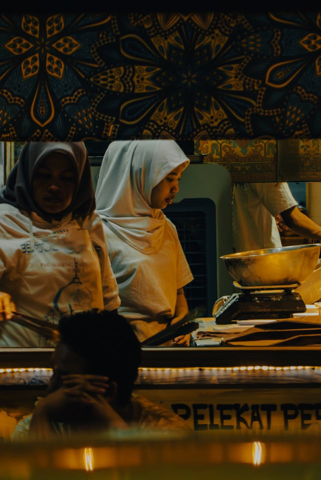 Two women in white headscarves work over pans at a food counter.