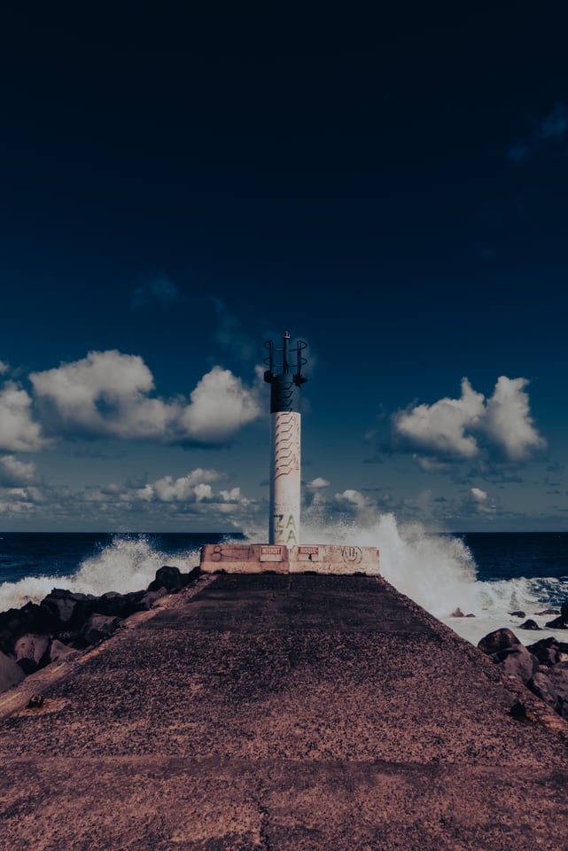 A wave breaks against the breakwater below a lighthouse tower.
