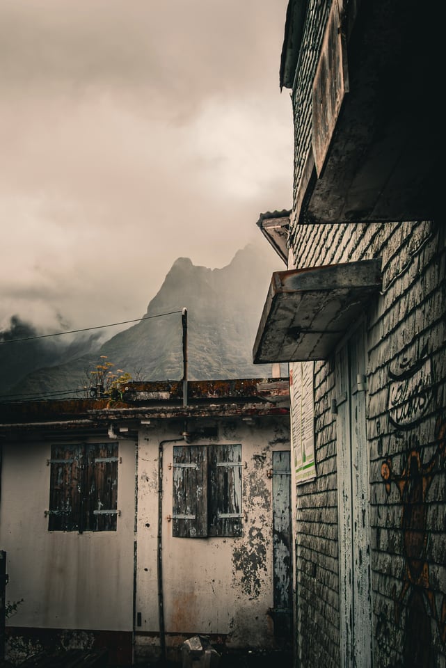 An abandoned house stands with mountains behind it in low cloud.
