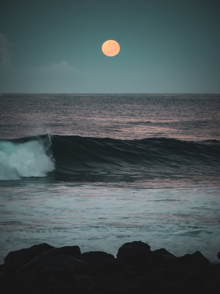 The moon stands above a breaking wave in the last light.