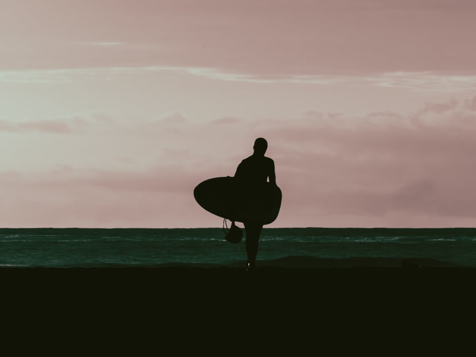 A surfer carries a board along the shoreline against a pale sky.