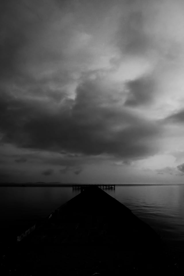 A dark pier point juts into a still sea under heavy cloud.