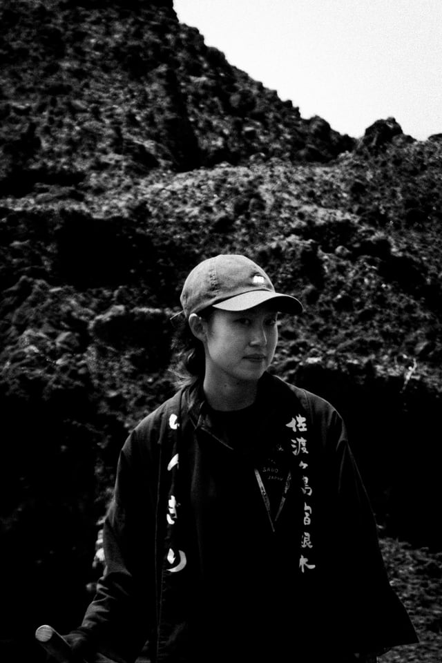 A woman in a cap stands against dark volcanic rock.