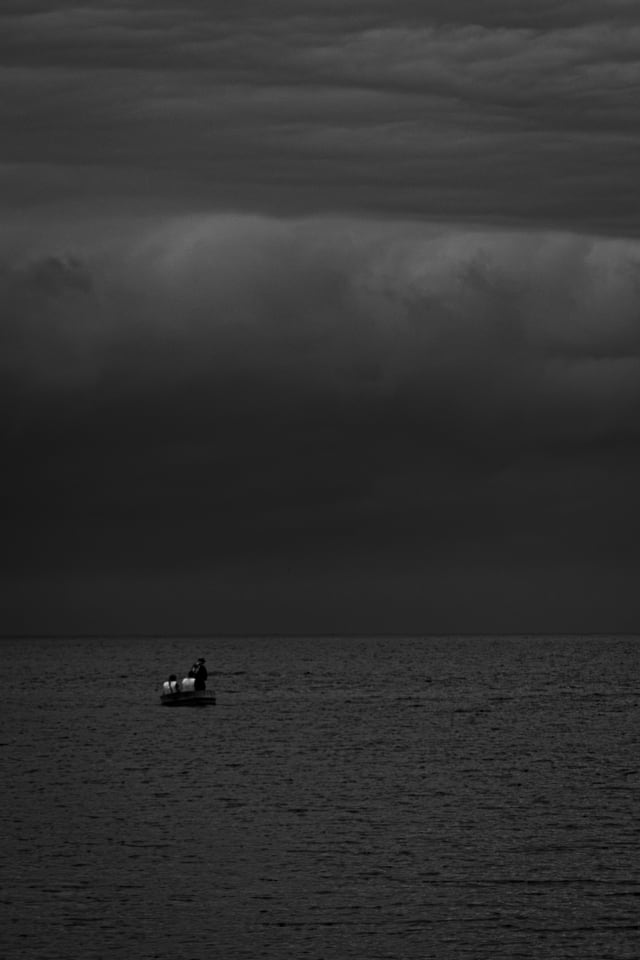A small boat sits alone on a wide sea beneath a heavy sky.