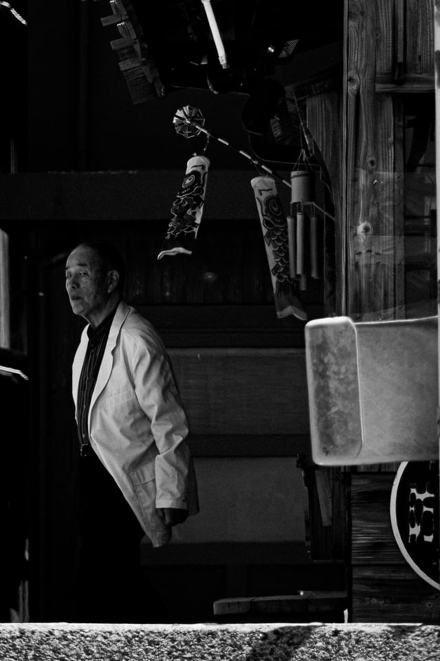 A man in a white coat stands in a dark shopfront hung with wind chimes.