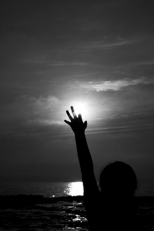 A silhouetted arm reaches up and cups the low sun above the water.