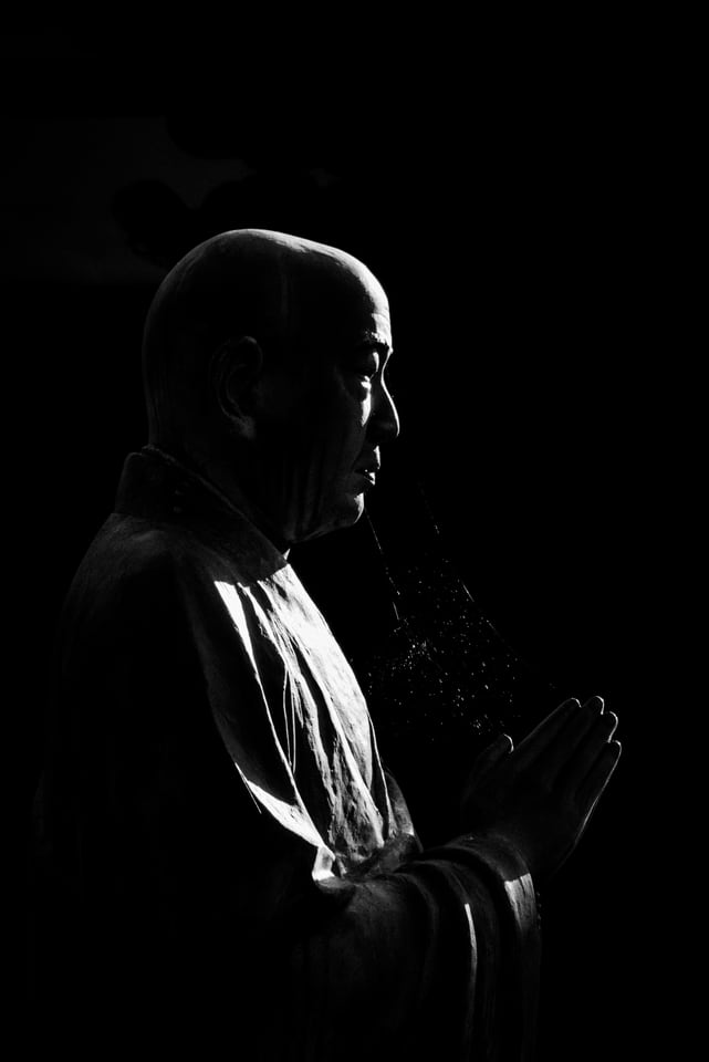 A person stands in profile with hands pressed together, lit from the side.