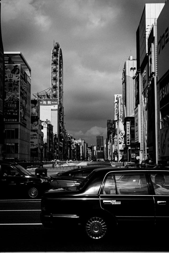 Taxis line a wide city street below signboards and a ferris wheel.