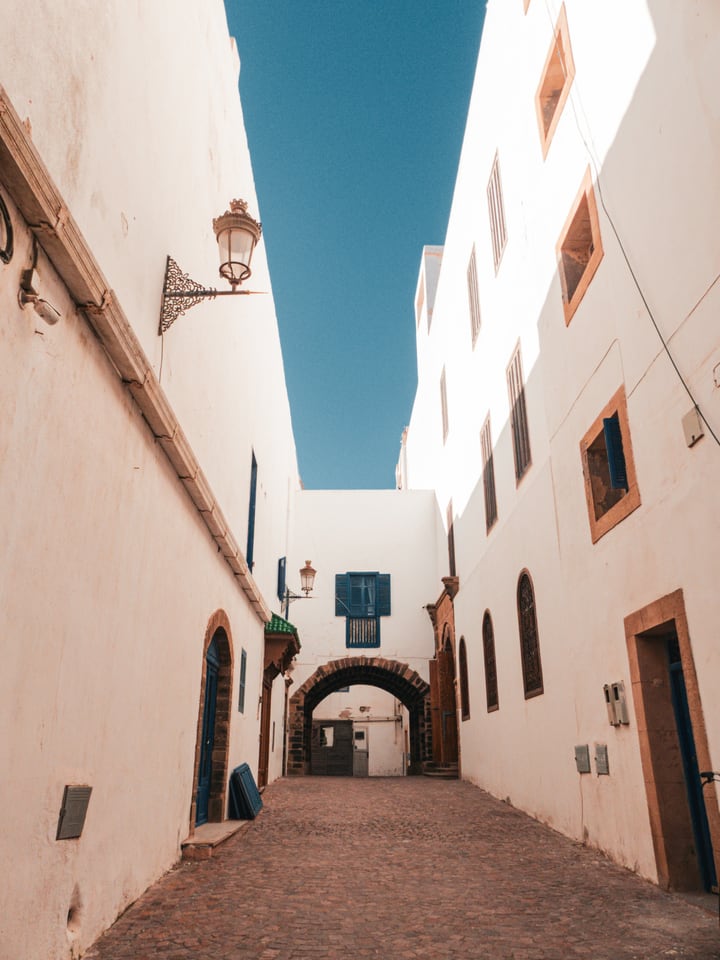 A narrow white lane leading to a stone archway beneath a strip of blue sky.