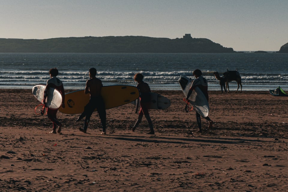 Surfers carry their boards along the sand below a headland.