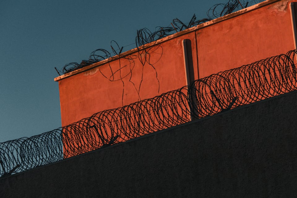 Coiled razor wire runs along the top of an orange wall against deep blue sky.