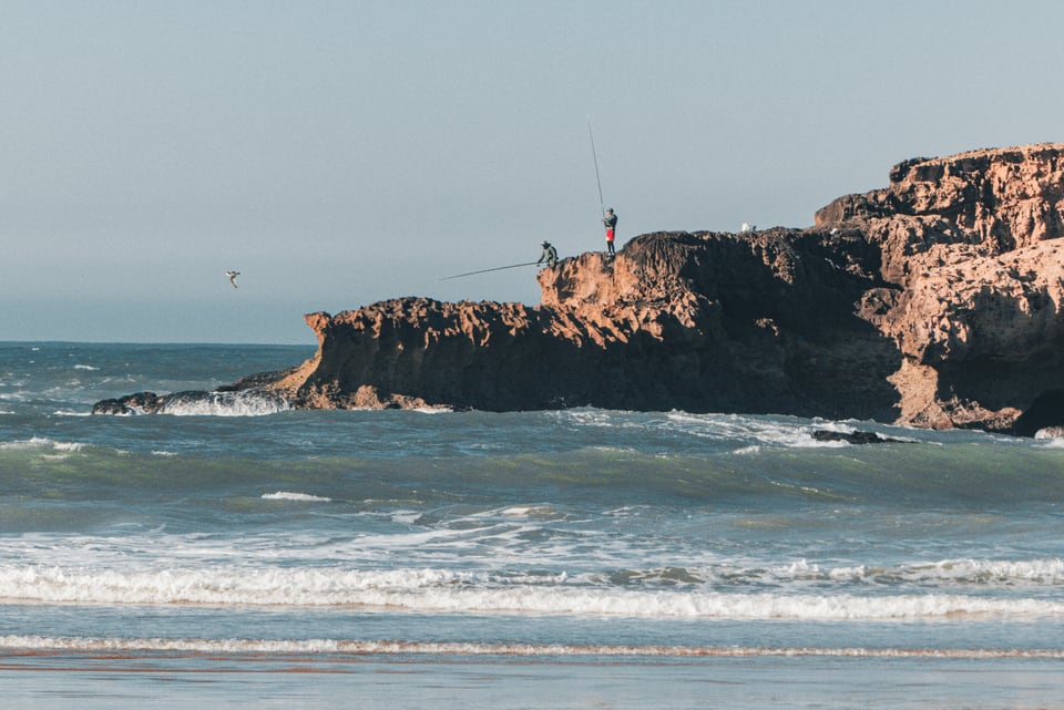 Waves break around a rocky outcrop with a small figure at its edge.
