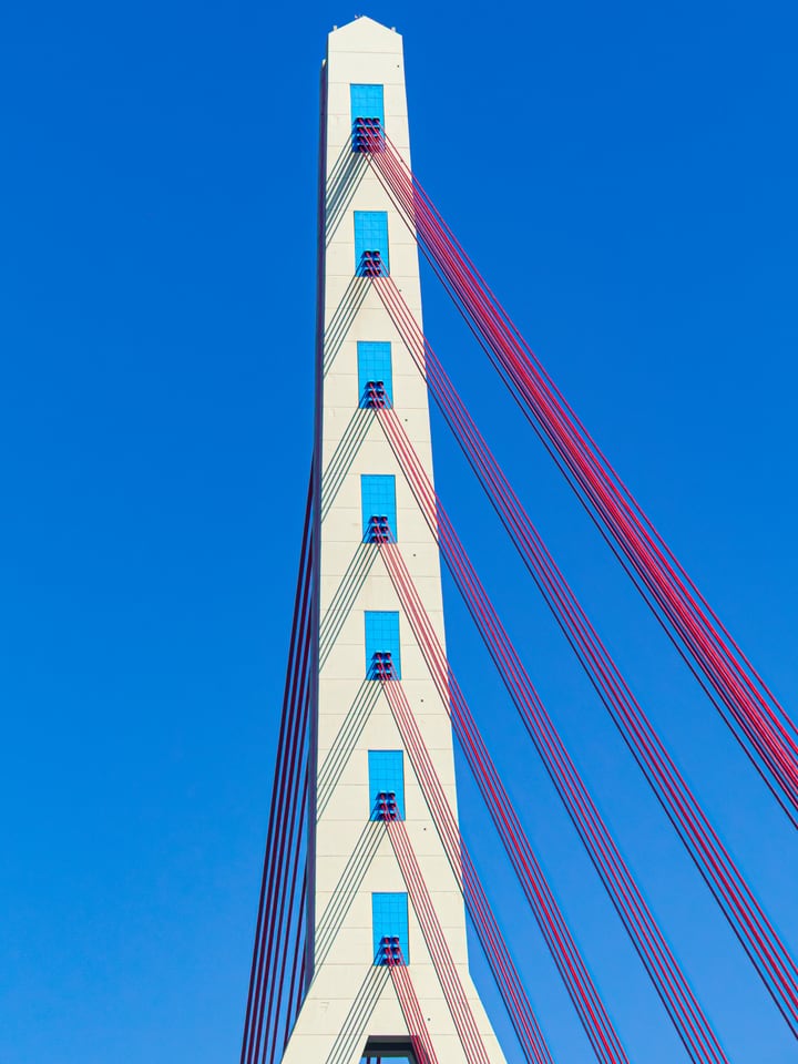 Red cables run down from the white pylon of a bridge into deep blue sky.