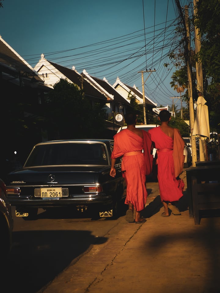 Two monks in orange robes walk along a Luang Prabang street past a black vintage Mercedes in low afternoon light.