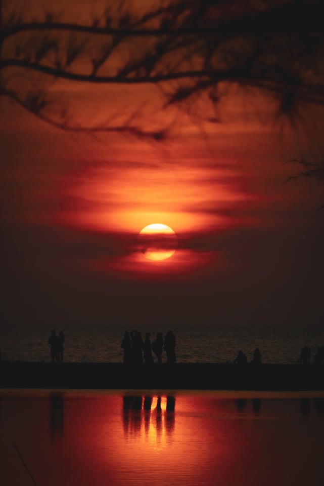 The sun drops behind cloud with figures on the shore below.