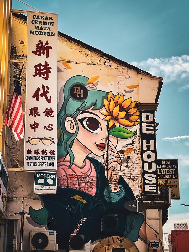 A painted girl holding a sunflower covers the gable end of a shophouse, beside a tall optician’s sign in Chinese and English and a Malaysian flag.