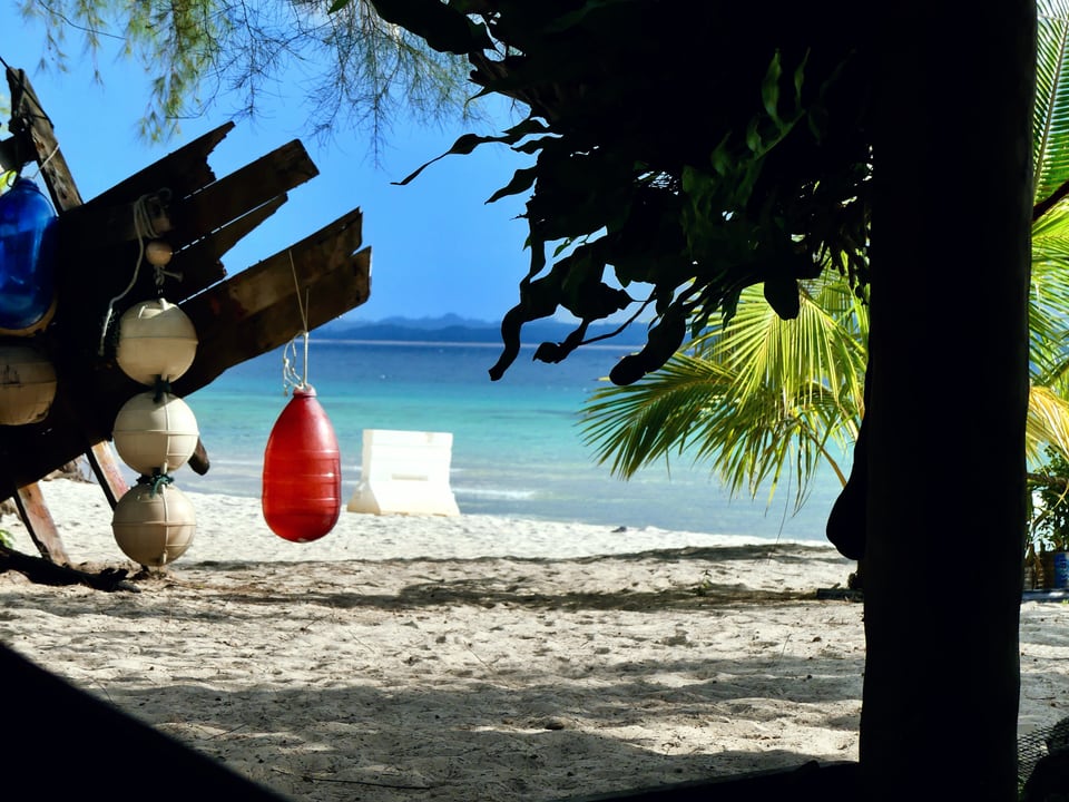Looking out from the shade of a shelter past hanging floats and palm fronds to bright sand, turquoise water and distant hills.