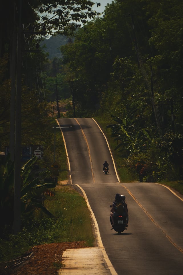 A road rises and falls through jungle with two scooters riding away into the distance.