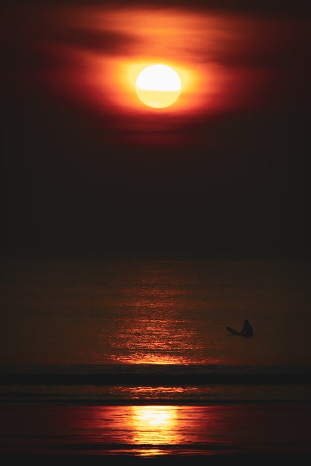 A large red sun stands low over the sea, with one small figure sitting on a board in the water beneath it.