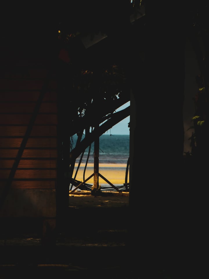 The beach and sea seen through a narrow opening between the dark walls and roof of a simple bungalow on Koh Phayam.