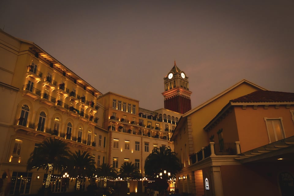 Illuminated Mediterranean-style buildings and a clock tower surround a quiet courtyard beneath a dark evening sky.